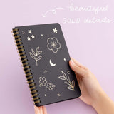 Tsuki Black Paper Ringbound Bullet Journal with beautiful gold details held in hands at an angle in lilac background