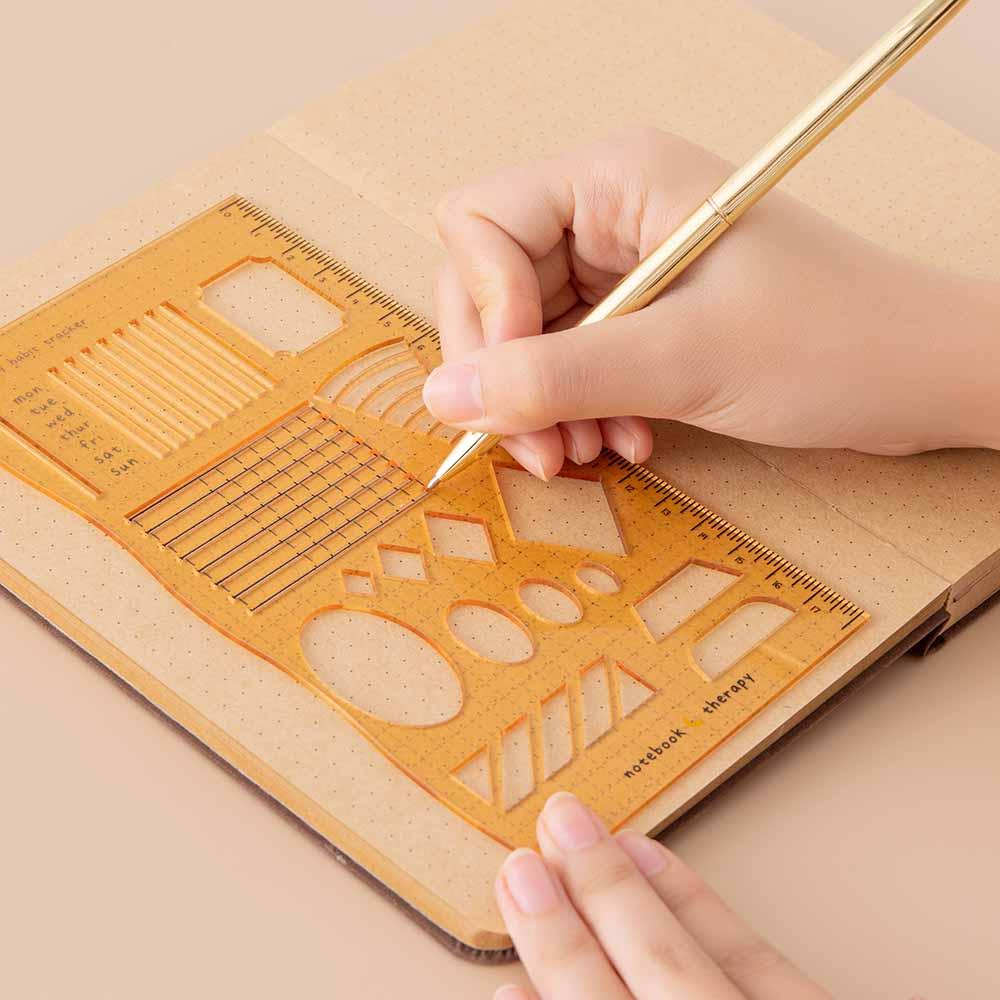 Tsuki Bullet Journal Stencil Set in neutral demonstrated on open bullet journal kraft paper page spread with pencil held in hands on beige background