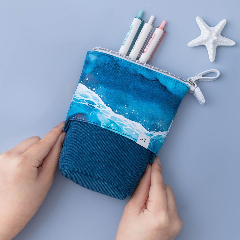 Tsuki Ocean Edition pop up standing pencil case in Ocean Blue held in hands at an angle with pens and starfish on blue background