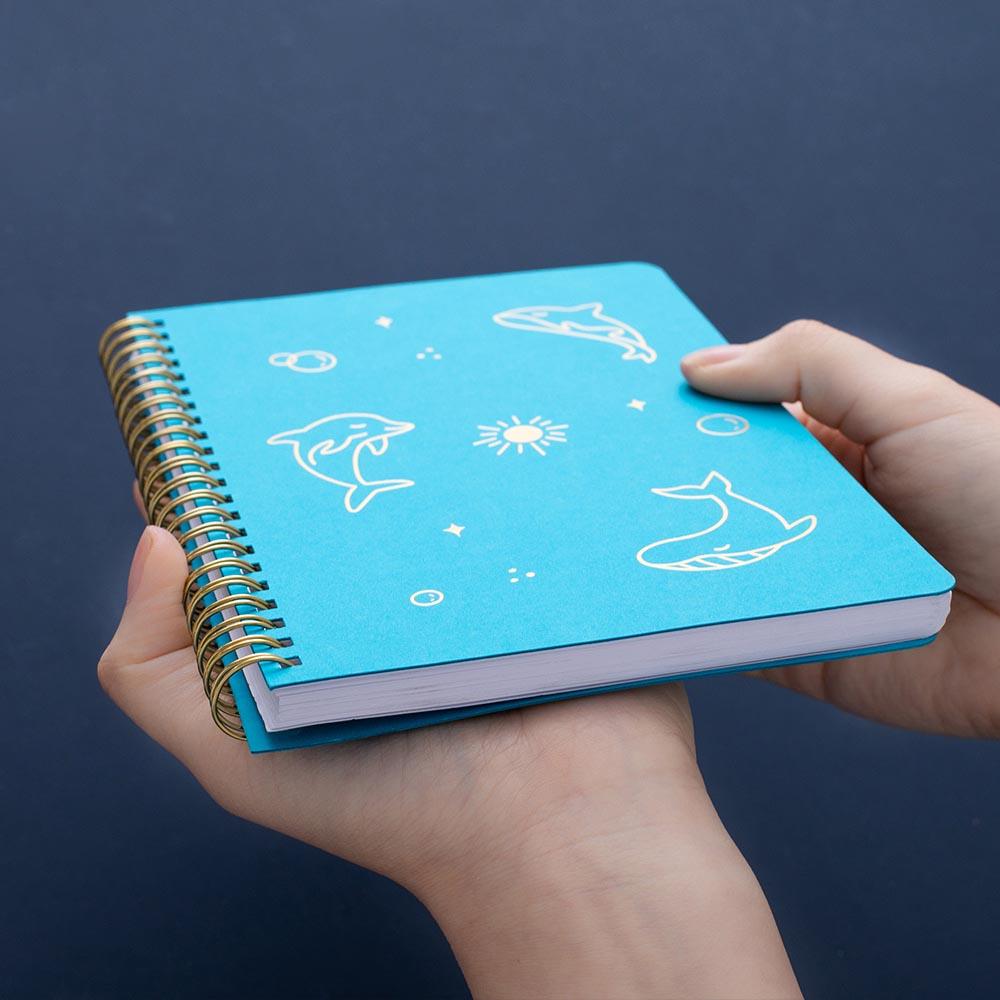 Tsuki Ocean Edition Ring Bound notebook in aqua blue held in hands at an angle on dark blue background