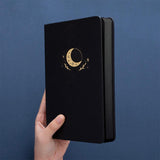 Tsuki Black Paper Limited Edition Hardcover Bullet Journal in Moonflower held in hands at spine angle in dark blue background