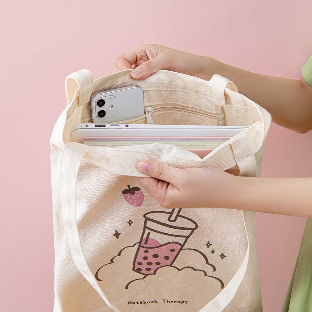 Close up of the inside of Tsuki ‘Ichigo’ Boba Tote Bag held in hands with laptop and mobile phone with inside zippable pocket in light pink background