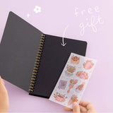 Open Tsuki Black Paper Ringbound Bullet Journal with free stickers sheet gift held in hands in lilac background