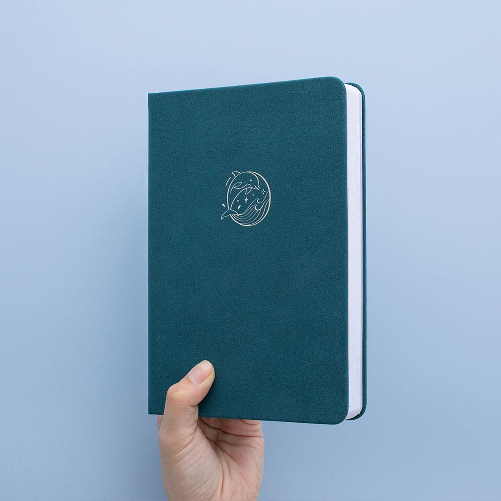 Tsuki sea green velvet Dolphin Days notebook held in hand at an angle in blue background