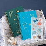 Tsuki Ocean Edition Ring Bound notebooks in aqua blue and deep teal with free sticker sheets and coral in basket on dark blue background
