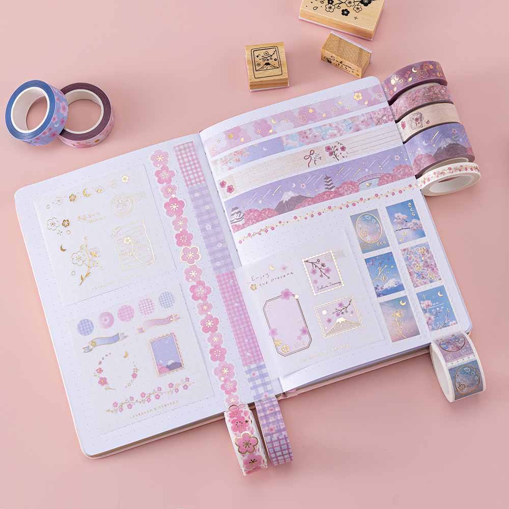 Tsuki ‘Sakura Journey’ Washi Tape Set rolled out on open Tsuki ‘Lunar Blossom’ Limited Edition Bullet Journal with Tsuki ‘Sakura Blossom’ Bullet Journal Stamp Set on light pink background