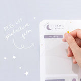 Close up of the front of Tsuki Reusable Weekly Planner with protective film being removed on light grey surface
