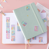 Tsuki Rainbow Pride Washi Tape on open notebook page with Tsuki Pastel Edition mint matcha bullet journal and postcard with bunting on light pink background