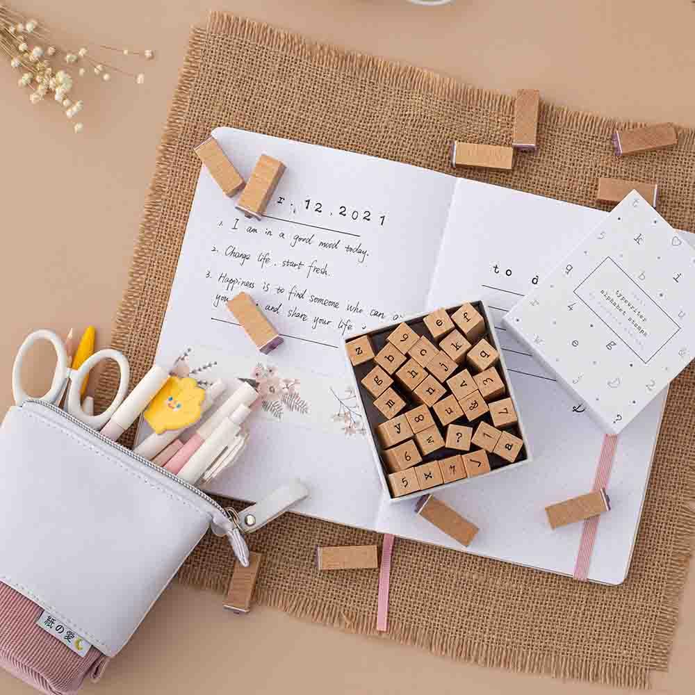 Tsuki Bullet Journal Typewriter Style Alphabet Stamps with eco-friendly gift box packaging and Tsuki sakura pink pop-up standing pencil case with open bullet journal and pens on hessian mat on beige background