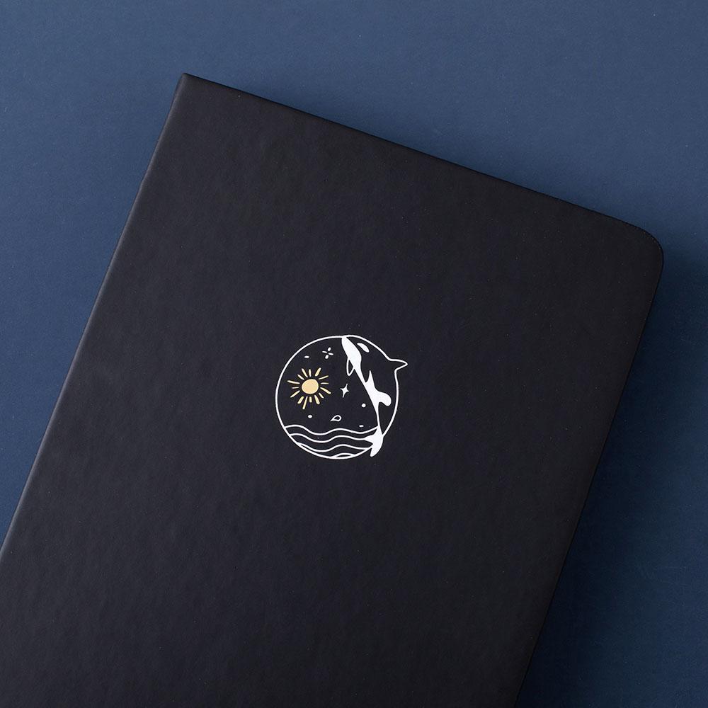 Close up of the front cover of deep black Tsuki Playful Orca limited edition bullet journal on dark blue background