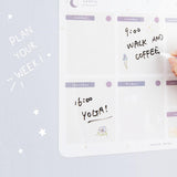 Close up of Tsuki Reusable Weekly Planner with dry erase marker held in hand on light grey surface