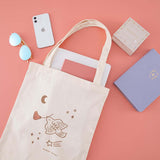 Tsuki ‘Moonflower’ Limited Edition Tote Bag with ‘Suzume’ Limited Edition Bullet Journal and laptop inside with ‘Full Bloom’ Limited Edition Notebook and ‘Moonflower’ Washi Tape Set with phone and sunglasses on coral pink background