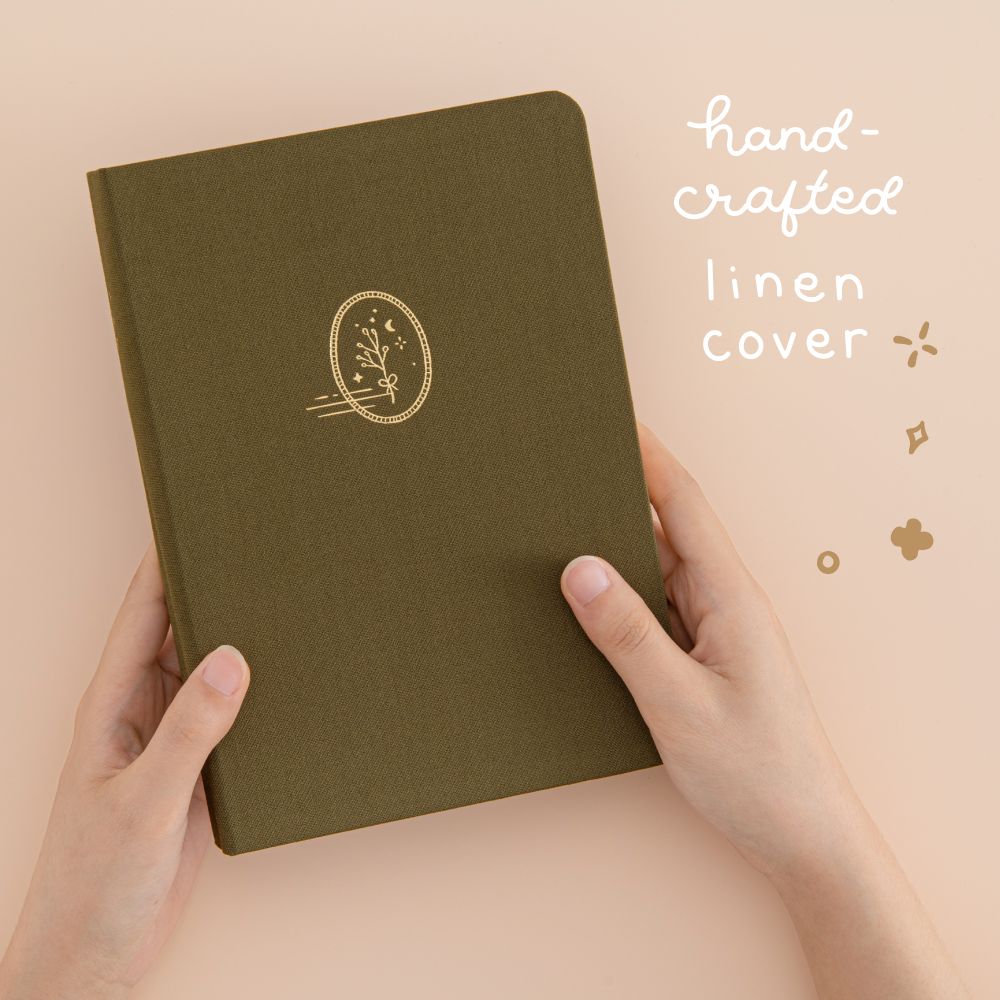 Hands holding green linen bullet journal with plant design with white lettering that says ‘hand-crafted linen cover’