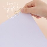 Hand putting ribbon shaped paperclip in gold on a bullet journal page with the words ‘free gift’ in white lettering