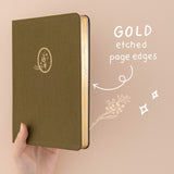 Hand holding up green linen bullet journal with gold gilded page edges