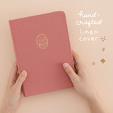 Hands holding pink linen bullet journal with cat design with white lettering that says ‘hand-crafted linen cover’