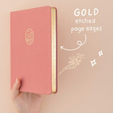 Hand holding up pink linen bullet journal with gold gilded page edges