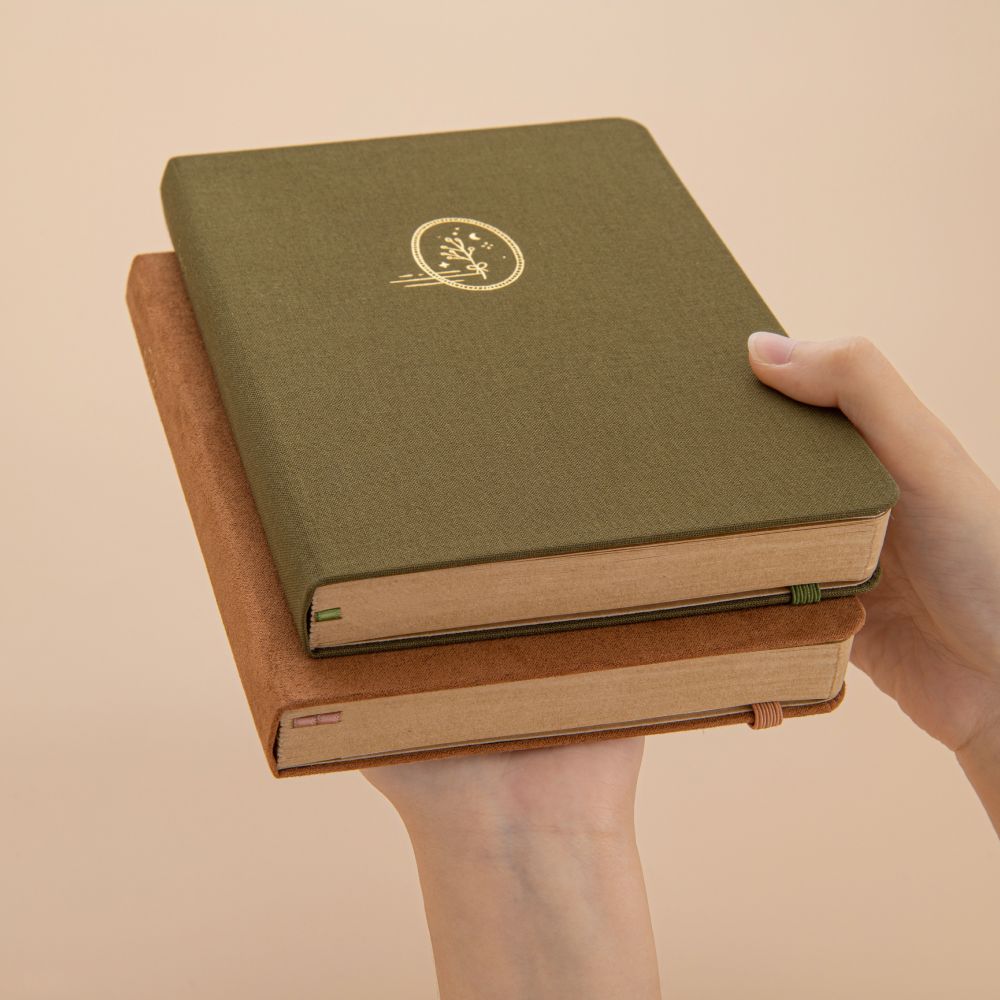 Hands holding Tsuki Mori green linen bullet journal and Kuma brown velvet bullet journal stacked on top of each other