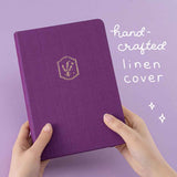 Tsuki ‘Enchanted Garden’ lavender foil design on purple linen bullet journal on purple background with white text saying ‘handcrafted linen cover’