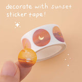 Sticker roll from Tsuki Golden Hour journal set and a close up of one sticker which is a picture of a sunset with gold foil details