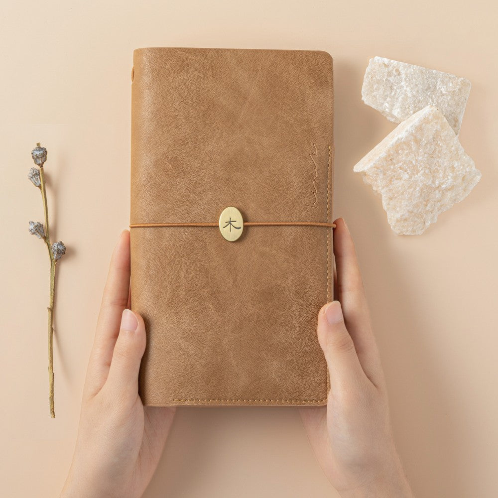 Hands holding the Hinoki - No.01 Travel Notebook, a brown leather notebook with an elastic band closure on a beige background with dried flowers and crystals on the side