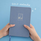 A pair of hands holding a blue bullet journal with a Tokyo design against a blue background and the text “gold details” written in white