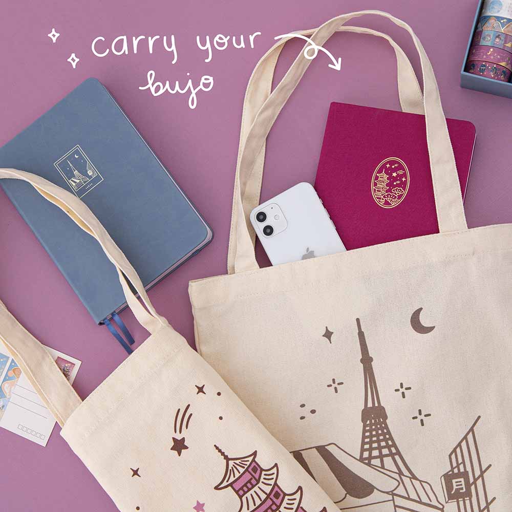 Tote bags on pink background with a notebook and phone peeking out of one canvas bag with the words “carry your bujo” in white