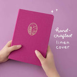 Two hands holding a fuchsia pink linen notebook with an illustrated temple in gold foil with the text “hand-crafted linen cover” written in white