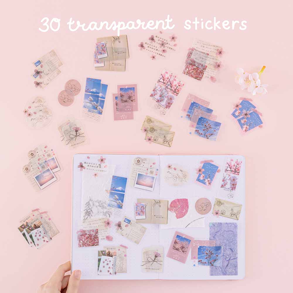 Thirty transparent stickers from Tsuki ‘Sakura Journey’ Scrapbooking Set on Tsuki ‘Sakura Journey’ Limited Edition Travel Notebook held in hand in pink background