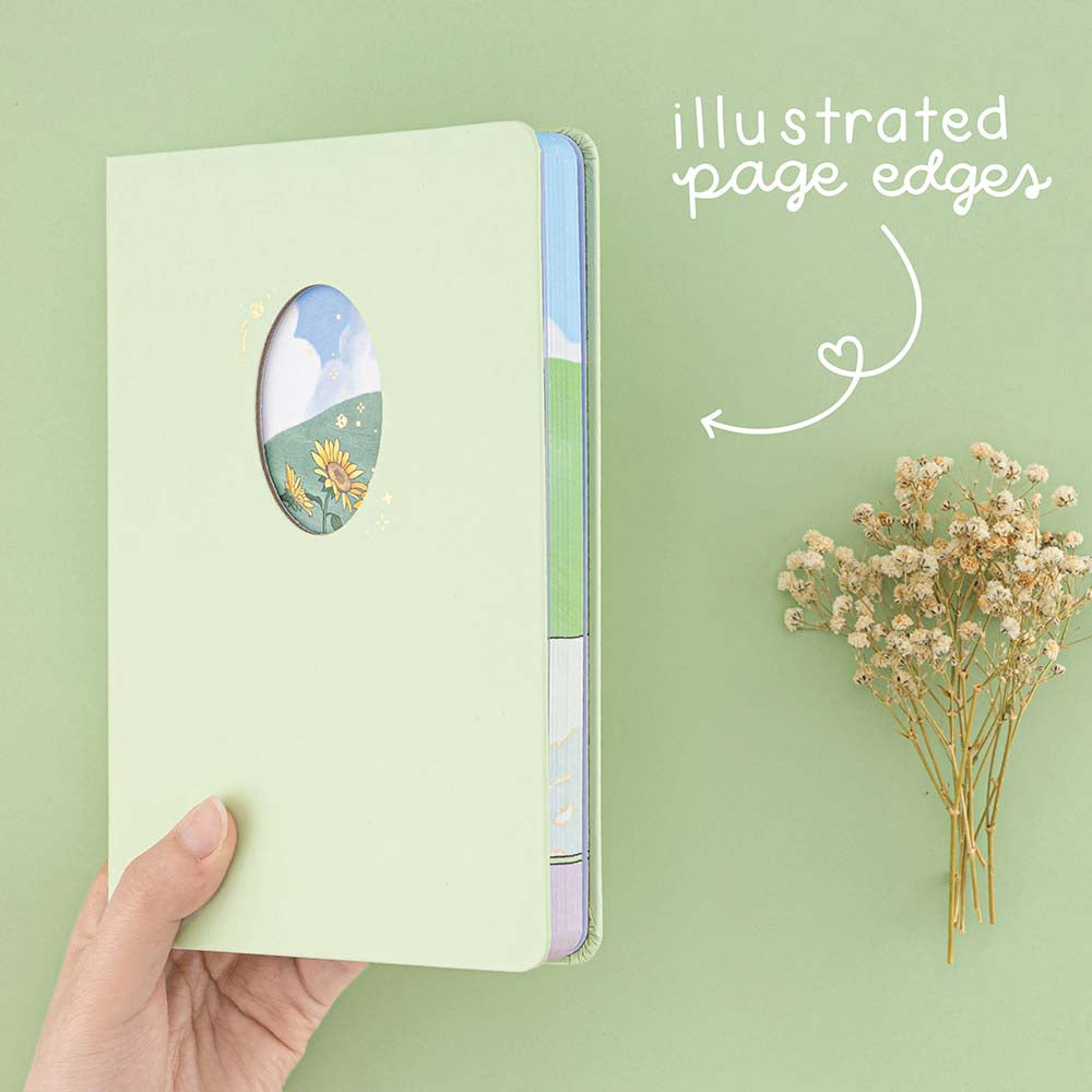 Hand holding Tsuki Four Seasons Summer Collectors Edition 2022 sage bullet journal notebook on sage green background with dried flower decoration and “illustrated page edges” in white handwritten text